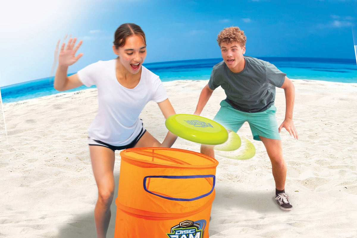 Two people playing with a bucket and frisbees on a beach.