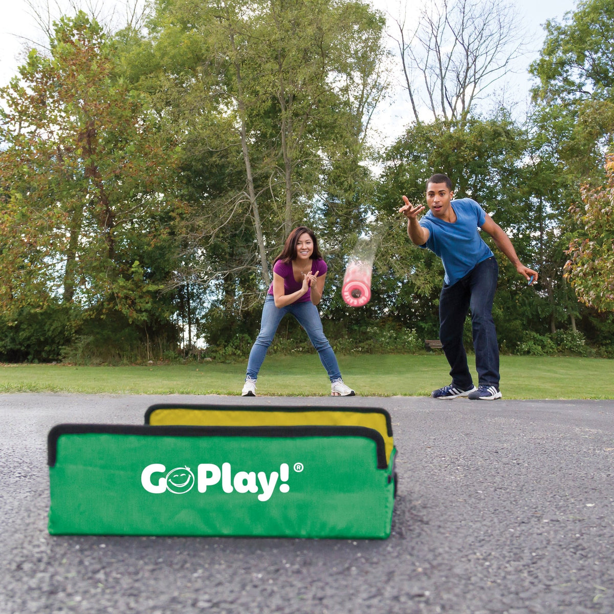 A man tossing a red washer towards a green Go Play! target outdoors while a woman watches.